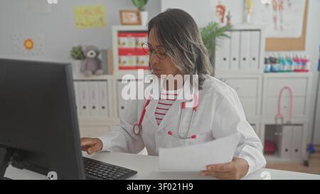 Medico ispanico donna in cappotto bianco tiene i documenti in mano e tipi su tastiera in un edificio medico indossando stetoscopio e occhiali; cura focalizzata. Foto Stock