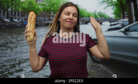 Donna che tiene la baguette e le onde mano sulla strada del canale di amsterdam mentre sorride in grande; viaggio divertente. Foto Stock