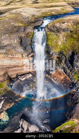 Vista aerea della cascata Haifoss che si getta su colonne di basalto con un vivace arcobaleno nella nebbia, circondato da un terreno verde muscoloso e scogliere rocciose Foto Stock