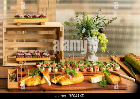 Un buffet da catering di crostini e antipasti infilzati è disposto su cassette di legno accanto a un vaso decorativo a forma di busto pieno di verde e uva Foto Stock