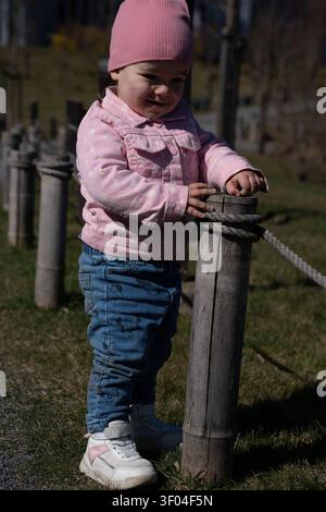 il bambino curioso con un berretto rosa e una giacca floreale si trova all'aperto con un palo in legno legato alla corda, inondato dalla luce soffusa del sole con un parco erboso Foto Stock