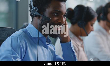 Ottimista e sorridente manager afroamericano nel call center consulente etnico addetto alla reception che parla in cuffia microfono digitando tastiera per computer Foto Stock