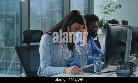 Donna d'affari caucasica stressata con mal di testa che lavora in call center operatori di team multietnici manager aziendali che parlano di linea telefonica con cuffie e auricolari Foto Stock