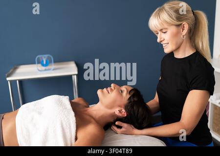 Terapeuta donna che esegue un massaggio seduto alla testa e al collo a un cliente maschile rilassato in una stanza di trattamento con parete blu, illuminazione soffusa e sedia coperta da asciugamano Foto Stock