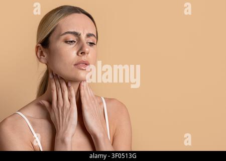 Giovane bella donna che tocca il collo con un'espressione preoccupata su uno sfondo studio beige Foto Stock