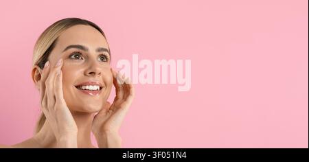 Donna sorridente che si tocca il viso e guarda joyosamente verso l'alto, mostrando una pelle liscia e radiosa Foto Stock