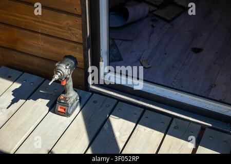 Avvitatore a percussione senza fili su terrazzo in legno all'ingresso di una casa privata, spazio di lavoro all'aperto soleggiato. Concetto: riparazione domestica fai-da-te Foto Stock