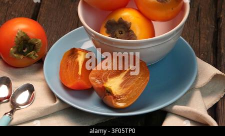 Persimoni interi e a fette di un arancione brillante sono disposti con arte in una ciotola di ceramica e su un piatto blu. Una luce laterale soffusa enfatizza la superficie liscia e cerata Foto Stock