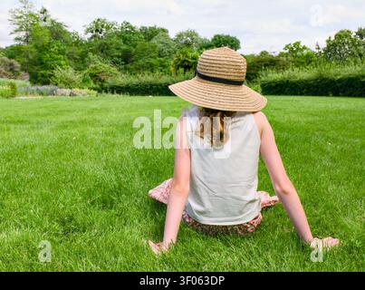 Una giovane donna, vista di spalle, è seduta sull'erba verde brillante di un parco, indossando un cappello di paglia e una gonna floreale Foto Stock