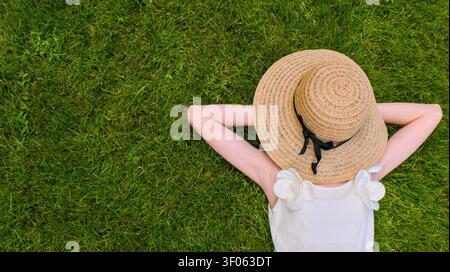 Una giovane donna, vista da una prospettiva dall'alto, giace su un prato verde vibrante con le mani dietro la testa, indossando un cappello di paglia e un top bianco. Foto Stock