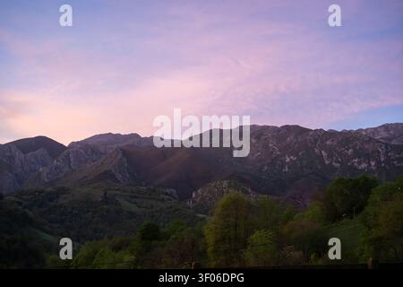 La luce soffusa del tramonto bagna la catena montuosa delle Asturie, con un tranquillo campo verde e colline boscose in primo piano sotto nuvole pastello, creando un'atmosfera unica Foto Stock