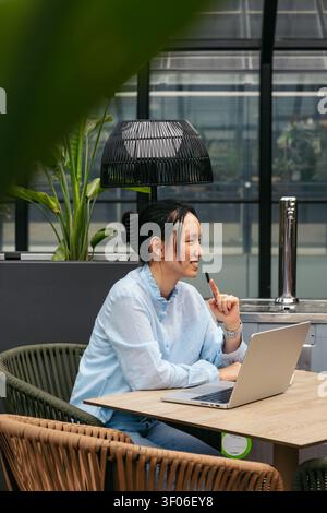 Una donna d'affari asiatica siede in uno spazio di lavoro all'aperto, sorridendo mentre tiene una penna e lavora sul suo computer portatile. L'ambiente moderno offre un lavoro rinfrescante Foto Stock