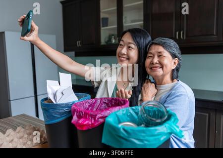 La madre asiatica sorridente e la figlia adulta fanno un selfie nella loro cucina mentre smistano carta e vetro in contenitori separati, promuovendo il riciclaggio e l'ecologia Foto Stock