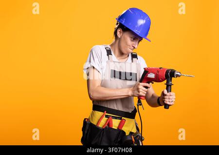 Donna bianca in piedi con sicurezza con avvitatore elettrico, indossa tuta protettiva e casco di sicurezza. Costruttrice esperta mostra capacità tecniche e prontezza a contribuire a progetti di costruzione. Foto Stock