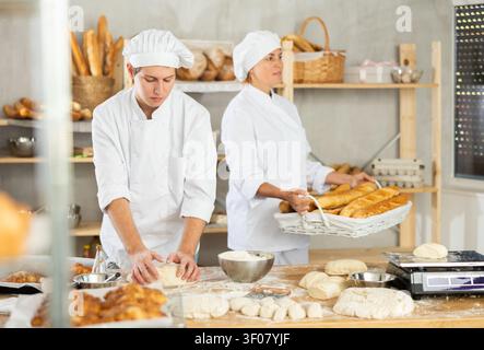 Fornai di successo qualificati in uniforme bianca che lavorano nel panificio. Il giovane che lavora con pezzi di impasto e la donna porta un cestino di prodotti da forno Foto Stock