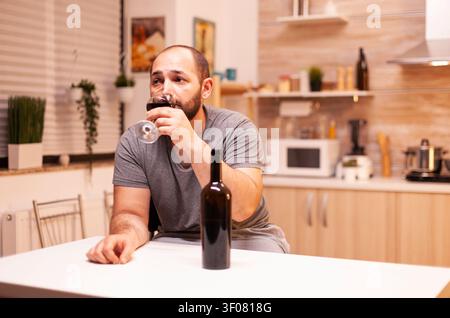 Uomo arrabbiato che beve vino rosso da solo al tavolo della cucina, beve da solo una bottiglia di vino rosso, seduto al tavolo in cucina. Persona infelice con malattia e ansia, stanca per l'alcolismo. Foto Stock