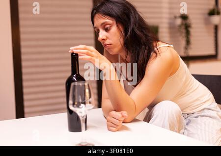 Donna triste che beve da sola in cucina Persona infelice che soffre di emicrania, depressione, malattia e ansia, si sente esausta con sintomi di vertigini e ha problemi di alcolismo. Foto Stock