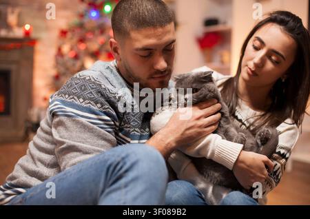 Coppia allegra gioca con il proprio gatto il giorno di Natale, godendosi un momento accogliente nella casa di vacanza con il proprio animale domestico, con decorazioni natalizie per la casa, illuminazione calda e interazione affettuosa della coppia. Foto Stock