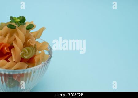 Pasta fusilli colorata in una ciotola di vetro isolata su uno sfondo azzurro, fotografia minimalista di cibo Foto Stock