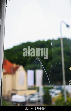Primo piano di una finestra incrinata con vetri rotti e un quartiere residenziale sfocato sullo sfondo, concetto di danno, vandalismo o riparazione. Foto Stock
