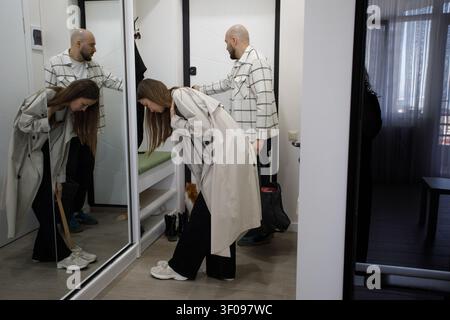 Una coppia è in piedi in un camerino. L'uomo guarda i vestiti appesi su una gruccia mentre la donna esamina il suo abbigliamento allo specchio. Stanno preparandosi Foto Stock