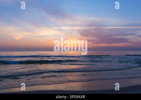 Un paesaggio marino sereno e grandangolare, cattura onde gentili che lambiscono una riva sabbiosa sotto un cielo dipinto di sfumature morbide di rosa, viola e blu Foto Stock