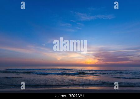 Un paesaggio marino sereno e grandangolare, cattura onde gentili che lambiscono una riva sabbiosa sotto un cielo dipinto di sfumature morbide di rosa, viola e blu Foto Stock