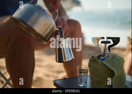 Il caffè mattutino viene versato da un bollitore di metallo in una pentola su una spiaggia sabbiosa accanto a un fornello da campeggio portatile, evocando relax, viaggio e cucina all'aperto Foto Stock