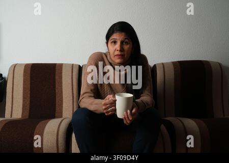 Donna seduta su un divano a righe, tiene una tazza bianca. Indossa un maglione marrone e pantaloni scuri. Il suo sguardo incontra direttamente la fotocamera. Una luce soffusa la mette in risalto Foto Stock