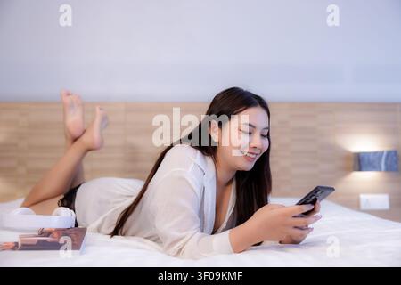 Una giovane donna in abiti casual da salotto sorride mentre usa il suo smartphone in un ambiente accogliente e contemporaneo da camera da letto. Foto Stock