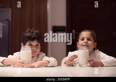 Due bambini del Medio Oriente siedono a un tavolo da cucina in una casa moderna, sorridendo mentre tengono in mano i bicchieri di latte durante una cena intima iftar per la celebrazione della famiglia Ramadan e Eid. Foto Stock