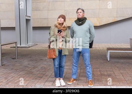Coppia senior in piedi sulla piazza lastricata di fronte a un edificio moderno che regge smartphone e borsa. Anziani, urbano, quadrato, telefono, borsa, sciarpa, berretto Foto Stock