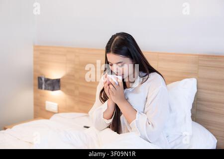 Una giovane donna è seduta a letto a rilassarsi con una tazza di caffè fumante al mattino, trasmettendo un senso di comfort e uno stile di vita tranquillo. Foto Stock