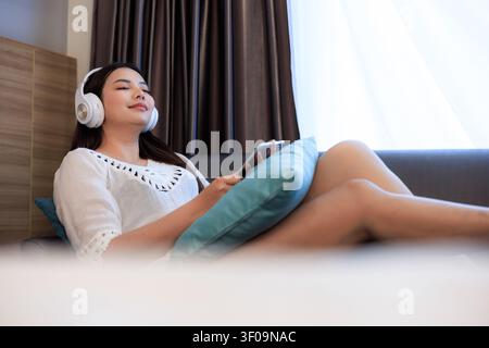 Una donna si gode il tempo libero sfogliando un tablet mentre indossa le cuffie su un divano che riflette uno stile di vita domestico sereno e accogliente. Foto Stock