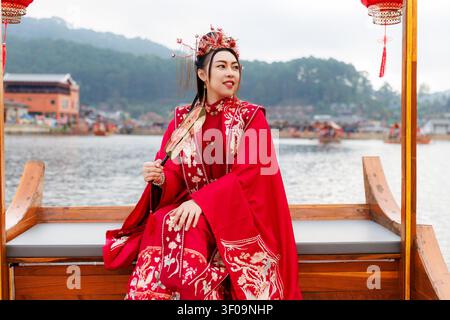 Una donna in tradizionale abito rosso hanfu tiene un ventaglio portatile mentre è seduta su una barca di legno a Ban Rak Thai a Mae Hong Son, Thailandia. Foto Stock