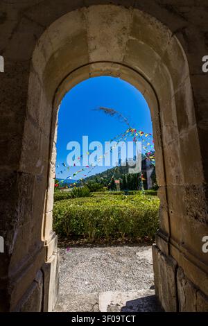 Incorniciato da un arco di pietra storico, un giardino cortile illuminato dal sole in Portogallo mostra bandierine colorate per le feste contro un cielo blu terso. Foto Stock