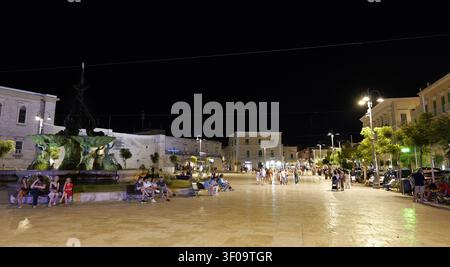 Persone che si godono una calda serata estiva nella piazza illuminata di Bisceglie, Puglia, Italia, con una fontana centrale. Italia, Puglia, Giovina Foto Stock