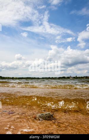 Riserva idrica del lago Lutskovlyany vicino a Hrodna, Bielorussia. Vista ravvicinata ad angolo basso di onde gentili che si infrangono su una riva sabbiosa sotto un cielo azzurro con un cl bianco Foto Stock
