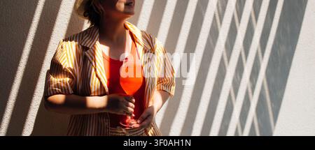 Donna in piedi contro un muro con ombre diagonali, tiene un bicchiere di bevanda arancione, vestita con una camicia a righe e un top rosso in uno spazio interno ben illuminato Foto Stock