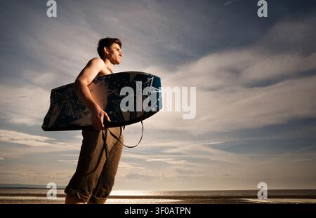 giovane uomo che tiene una bodyboard in piedi su una spiaggia guardando verso il mare Foto Stock