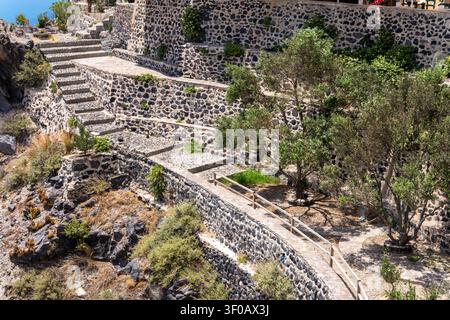 Scale di ciottoli sinuose e muri di sostegno terrazzati in pietra vulcanica. Alberi di ulivo verdi e vegetazione mediterranea rada crescono su un h soleggiato Foto Stock