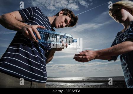 giovane uomo che lava le mani di una donna versando acqua da una bottiglia di plastica su una spiaggia Foto Stock