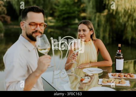 Un uomo e una donna si siedono a un tavolo di vetro vicino a un corpo d'acqua. Tengono i bicchieri da vino e si guardano a vicenda. Sul tavolo sono disponibili vari piatti. È e. Foto Stock
