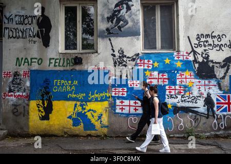 Tbilisi, Georgia. 7 luglio 2026. Le donne passano davanti a un muro coperto di Street art politica e graffiti, tra cui bandiere ucraine e georgiane a Tbilisi, Georgia, il 7 luglio 2026. (Foto di Moe Zoyari/Sipa USA) credito: SIPA USA/Alamy Live News Foto Stock