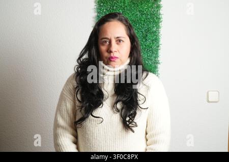 Donna in piedi davanti a una siepe artificiale verde. Indossa un maglione a collo alto color crema. I capelli sono lunghi, scuri e mossi. Il muro dietro di lei è semplice Foto Stock