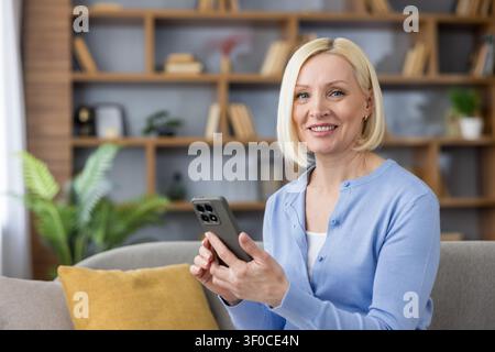 Donna bionda di mezza età seduta su un divano, utilizza felicemente uno smartphone per comunicare e connettersi, godendosi uno stile di vita moderno nel suo ambiente domestico confortevole Foto Stock