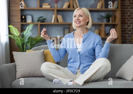 Donna matura seduta a gambe incrociate su un divano, che pratica la meditazione con le mani in una gestualità mudra, trovando la pace interiore e promuovendo il benessere nel suo soggiorno confortevole Foto Stock