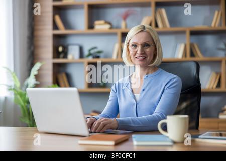 Donna professionista matura seduta a una scrivania moderna, sorride alla telecamera mentre digita su un laptop, incarnando uno stile di lavoro remoto di successo e sicuro di sé Foto Stock
