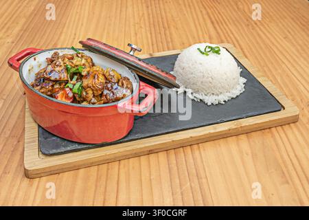 Pollo brasato in salsa ricca servito in pentola di ghisa rossa con riso bianco al vapore su tavola di ardesia Foto Stock