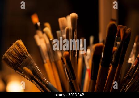 I pennelli dell'artista riposano in un laboratorio artistico, circondati da opere su tela e strumenti creativi contemporanei per la pittura e la pratica delle belle arti. Foto Stock